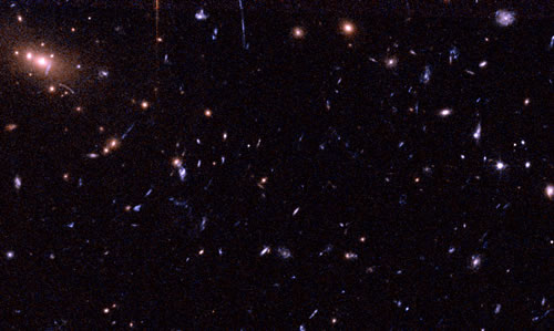This image shows a galaxy cluster (RXJ1226.9+3332) at a great distance (redshift 0.89). Yellowish/orange objects are massive galaxies in the cluster, while faint blue elongated shapes are distant galaxies stretched by the cluster's gravity.
